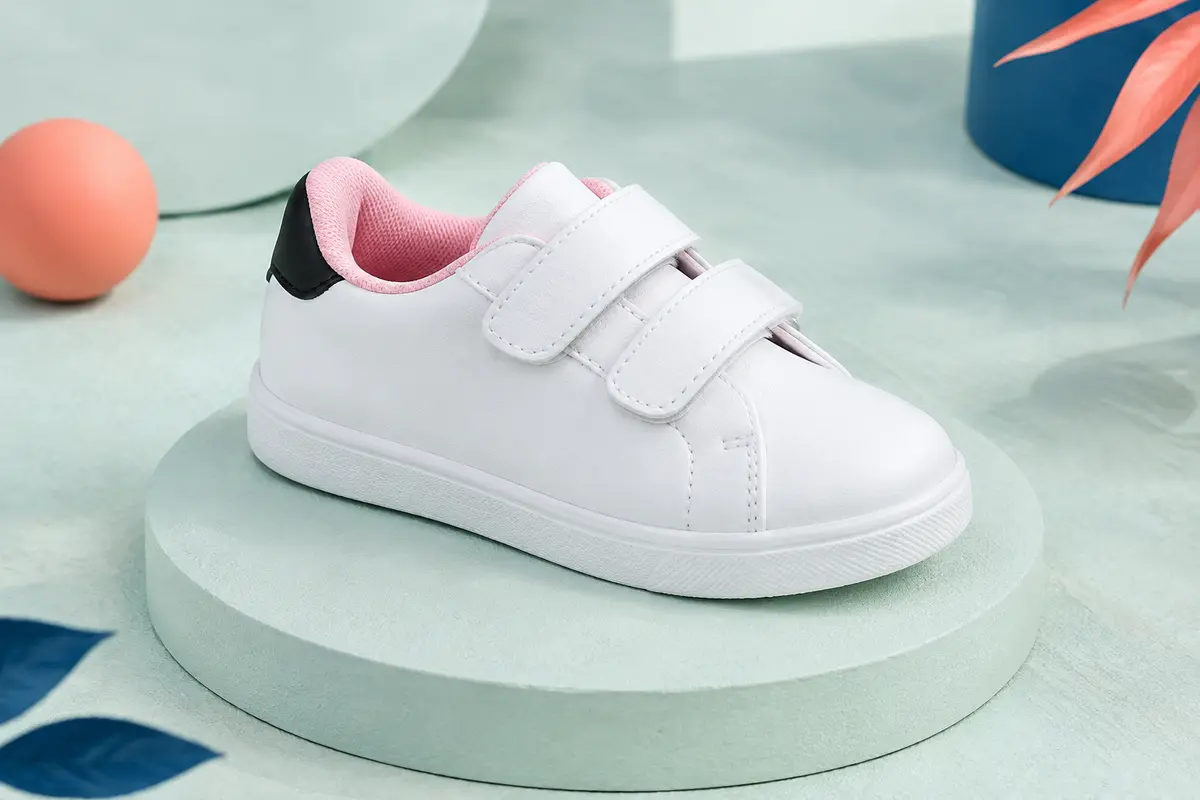 Basket basse enfant blanche à brides autoagrippantes, doublure rose et talon noir, posée sur un socle mat.