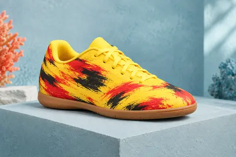 Chaussure de futsal enfant jaune à motif abstrait rouge et noir, semelle plate couleur gomme, posée sur un socle mat.