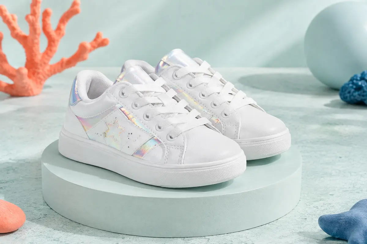 Baskets basses Adidas blanches à lacets avec détails irisés, posées sur un socle mat dans un décor pâle.
