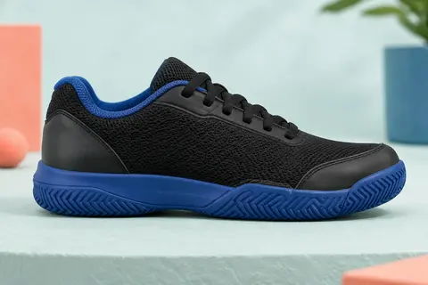 Chaussure de tennis junior noire à col rembourré et semelle à chevrons bleu profond, posée de profil sur un socle mat.
