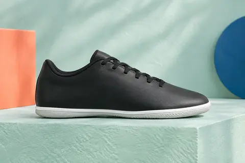 Chaussure de futsal junior noire unie à semelle plate claire, posée de profil sur un socle mat.