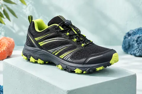 Chaussure junior de trail basse, sombre avec accents citron, posée sur un socle dans un décor lumineux